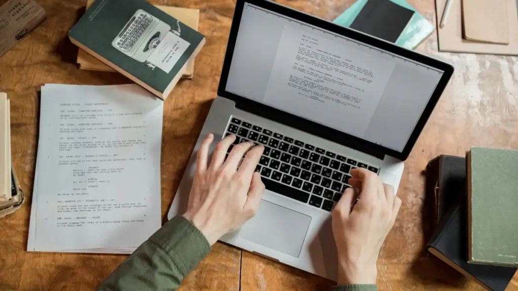 Copywriting Blog Image: Laptop, Hands, and Books Hands typing on a laptop keyboard with books and papers nearby, illustrating copywriting work
